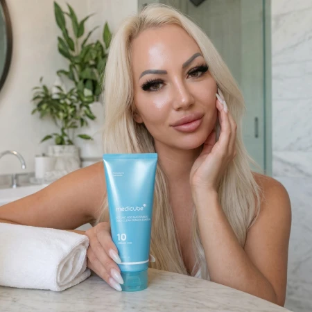 Kobieta o promiennej cerze trzymająca w dłoni opakowanie pianki do mycia twarzy Medicube Azelaic Acid Niacinamide 120g w jasnej łazience.